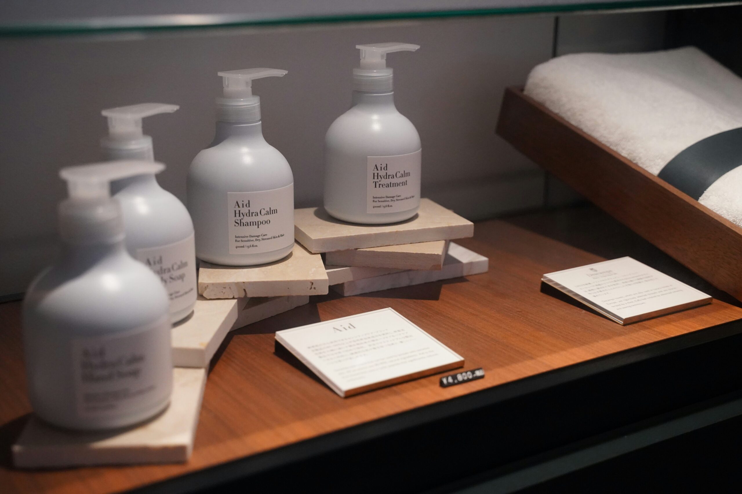 A set of Aid Hydra Calm shampoo, treatment, and body soap bottles displayed on stacked stone blocks in a store, next to informational cards and a folded towel.